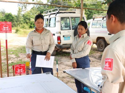 Task supervisor, Ly Sreyneang and unit commander, Pat Liv, stand behind tables with mine warning signs behind them