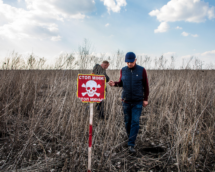 Two men walk in a agricultural field with a red mine warning sign