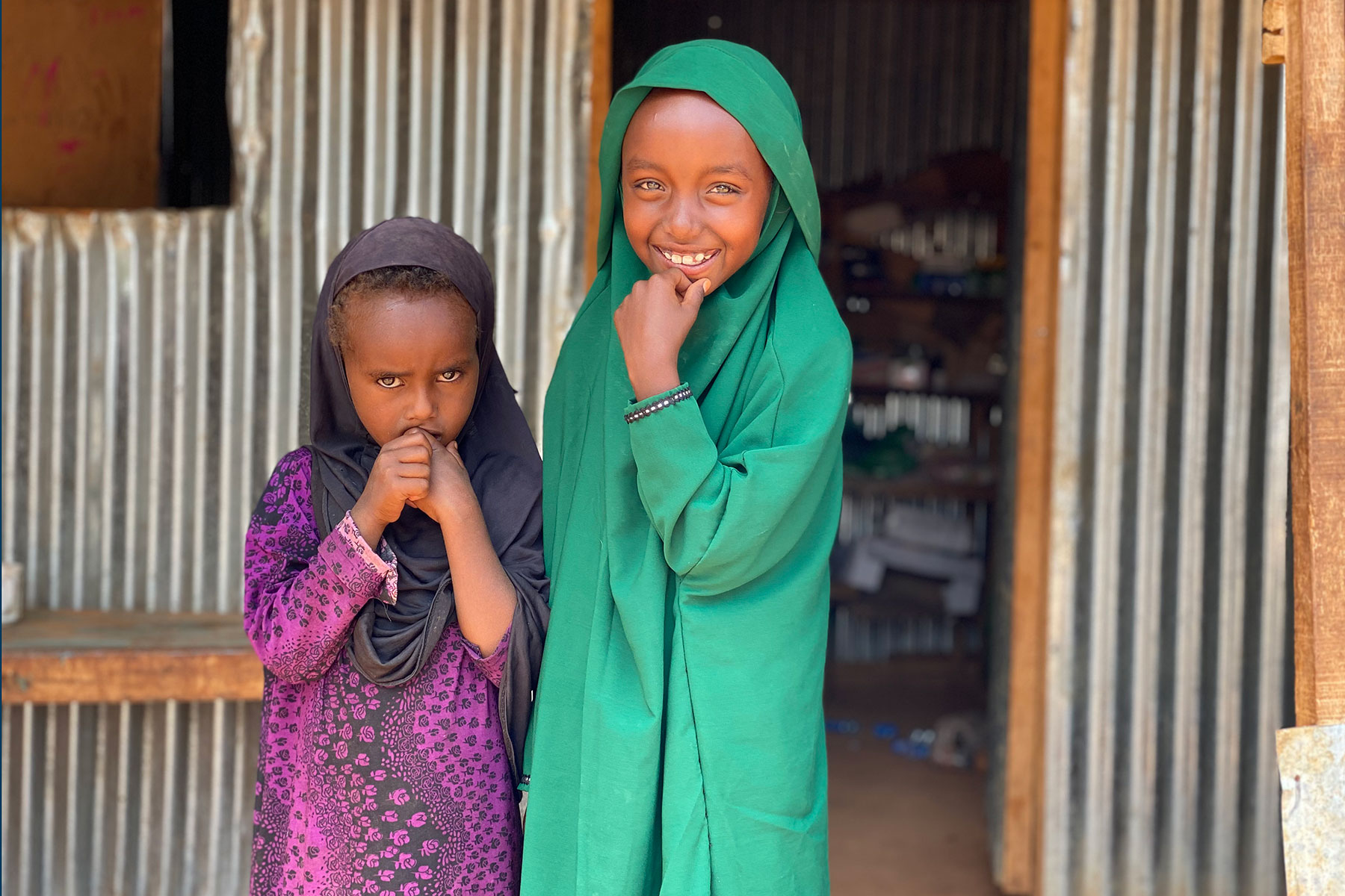 Two young girls stand outside a room made from metal sheets