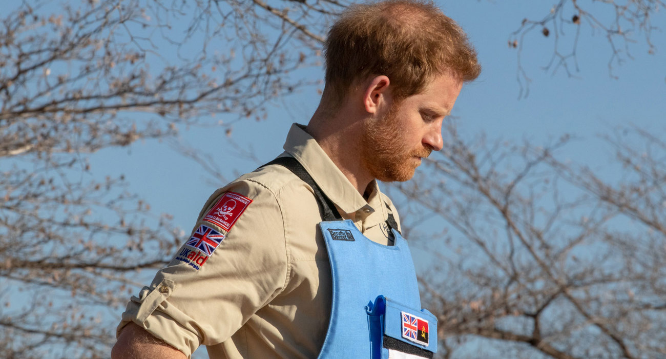 Prince Harry stands in a minefield in Angola wearing HALO uniform and a protective vest displaying the UK flag
