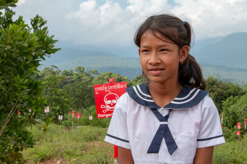 A girl sits in front of a danger mines sign in a field in Cambodia