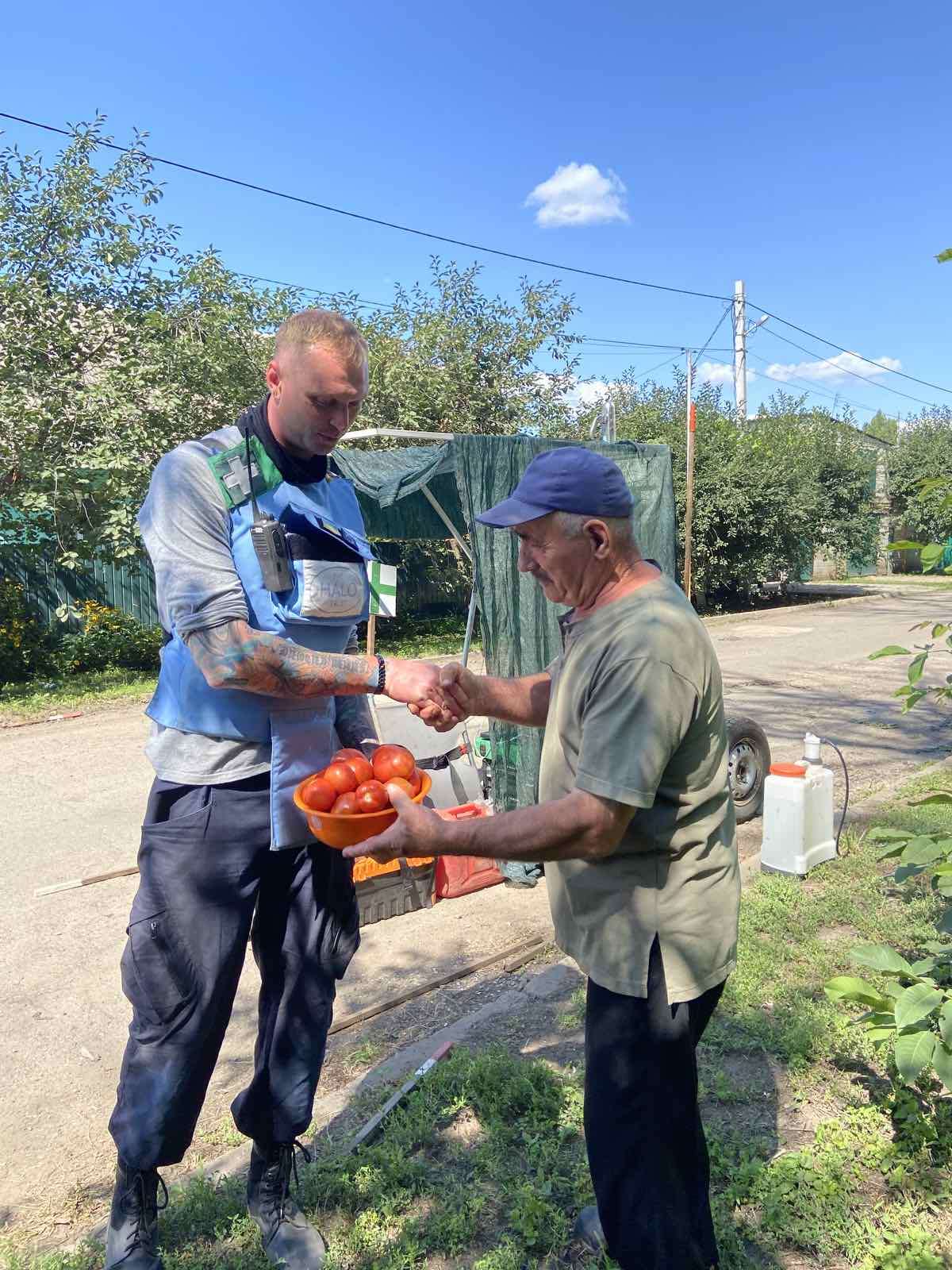 A HALO deminer in PPE shakes hands with a Ukrainian farmer holding a small basket of tomatoes.