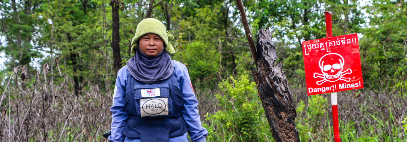 A deminer in Cambodia holds a detector and visor while walking through a rural minefield next to a red danger mines sign