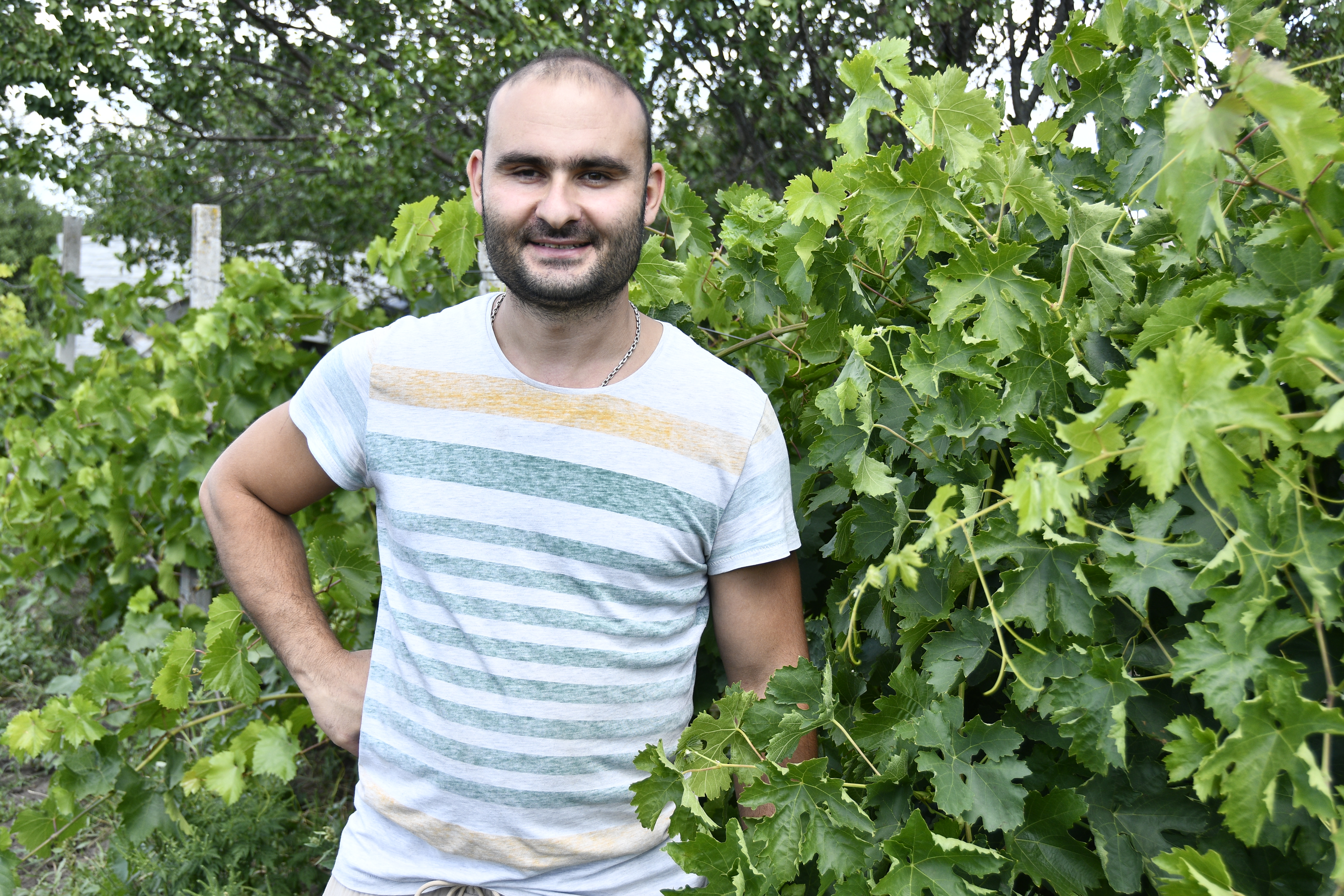 A man in a striped t-shirt stands smiling in front of a leafy bush.