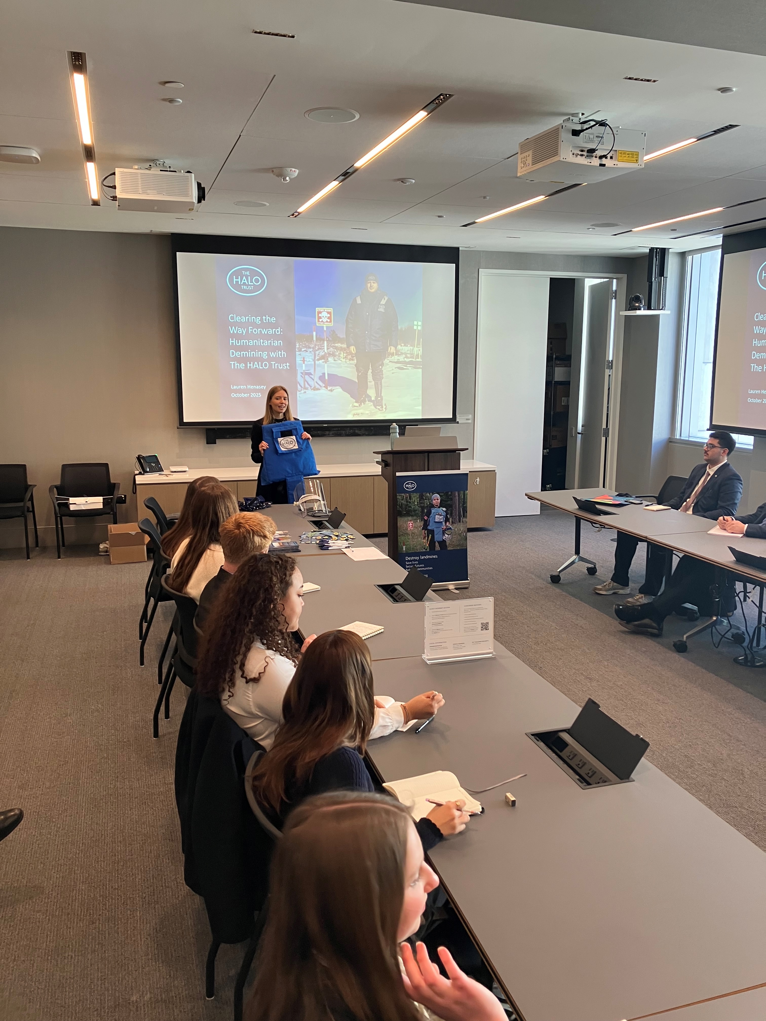 Lauren, a HALO team member, holds up a blue demining vest while speaking to university students.