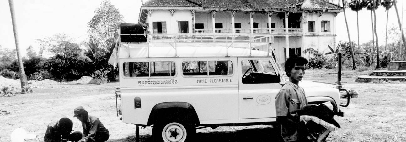 A black and white image of a HALO vehicle in front of a house in Cambodia, with HALO Trust staff 