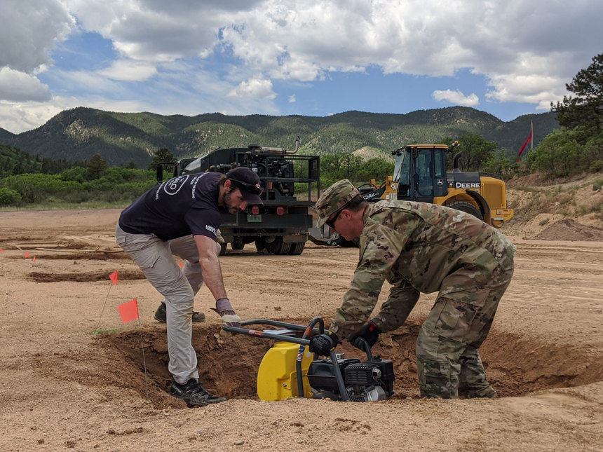 A HALO and a USAFA staff member lay a piece of machinery in a hole to test devices designed to detect landmines and explosives