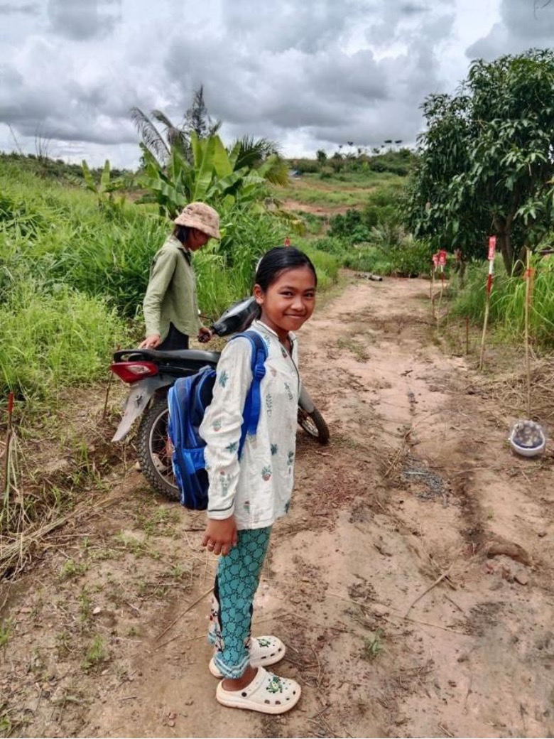 A young girl wearing a backpack stands in front of a path through high grasses marked by minefield signs on one side.