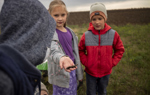 Three young children play together in a field. A girl stands in the middle, showing a small brown object to the camera.