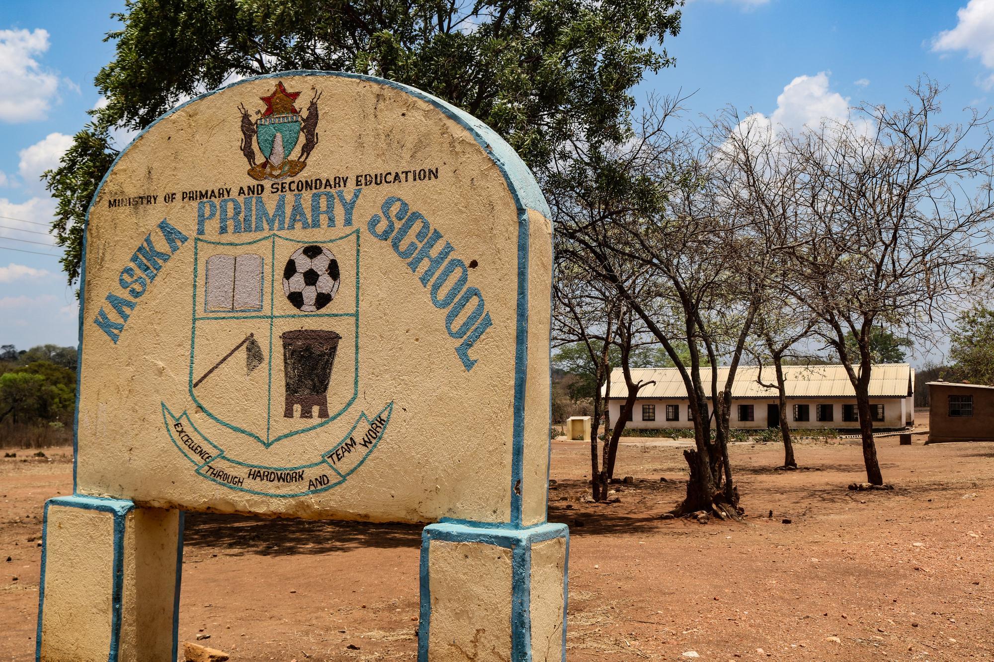 A sign for Kasika Primary School along with the school's crest stands outside of a modest school house