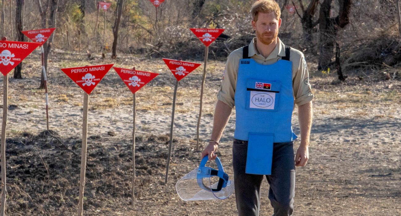 Prince Harry walks through a minefield in Angola, wearing PPE and holding a visor next to red danger signs