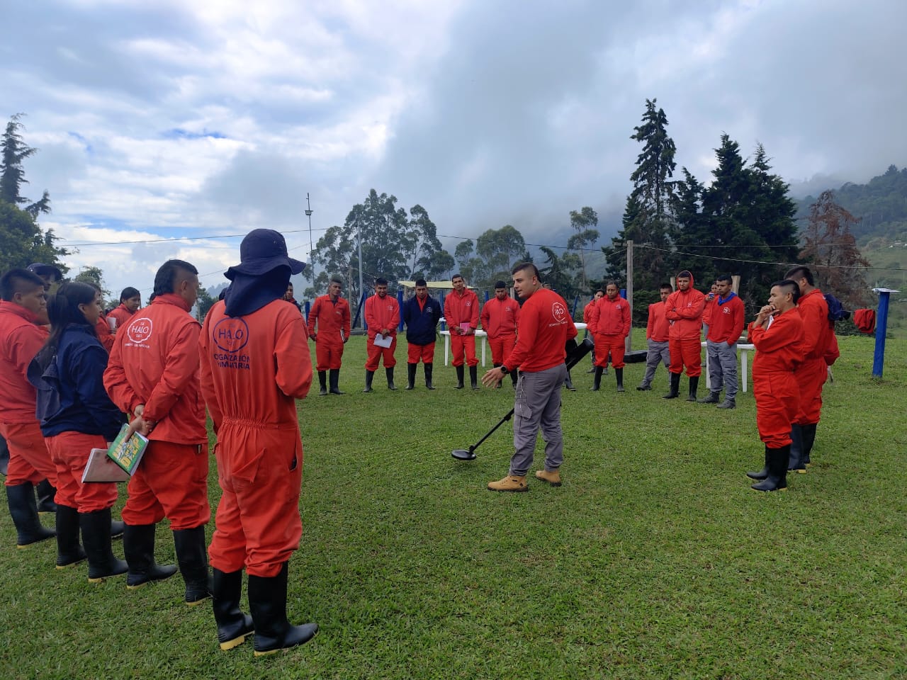 Deminer John Alexander stands with a metal detector in the middle of a semicircle of HALO team members, all in red HALO clothing.