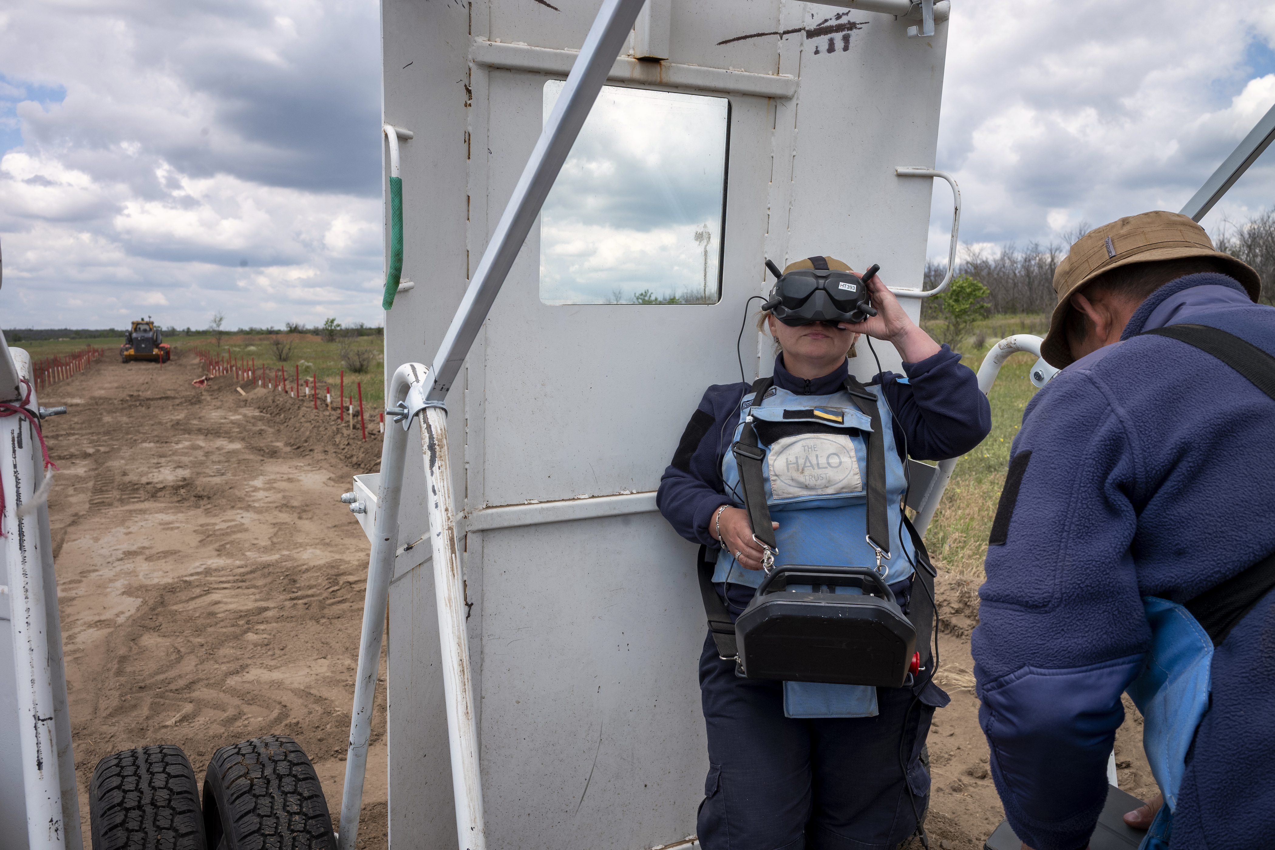 A woman in a HALO Trust vest operates a large remote control, controlling a large vehicle from a distance. 