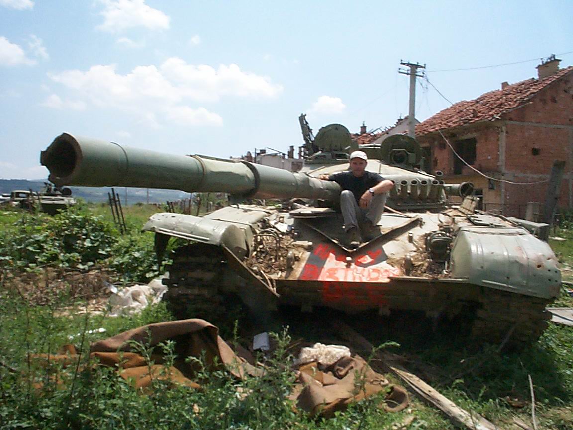 A man sits on an old abandoned tank in the kosovan countryside, surrounded by rubble and damaged houses