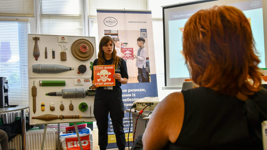 A HALO staff member holding a danger sign in front of demonstration explosives in a risk education class