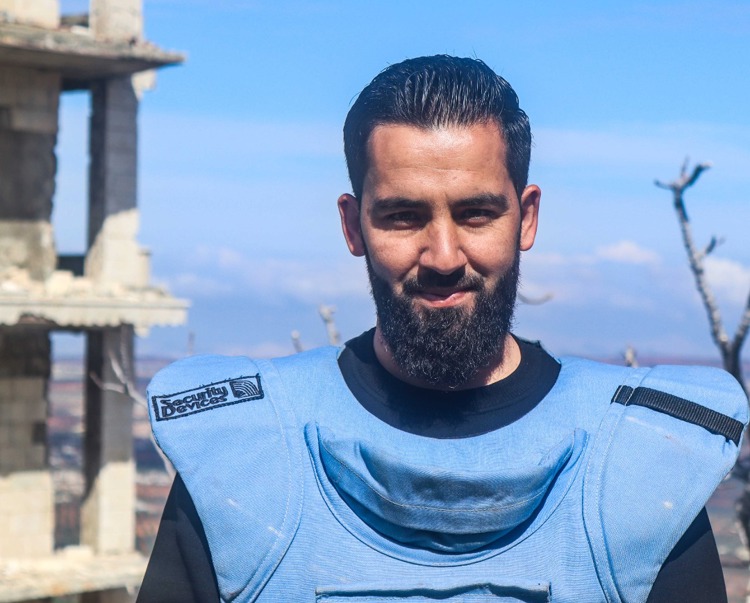 Farooq Al-Mustafa, stands in PPE in front of a damaged building in Syria