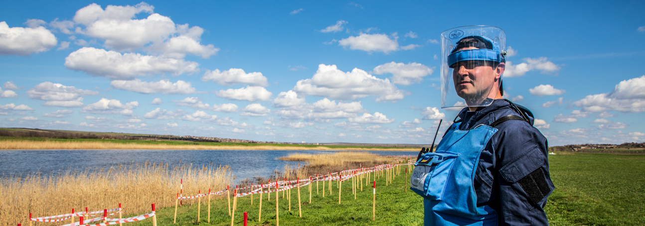 A HALO Trust deminer stands in a rural minefield next to a lake, with contamination clearly marked in front of him