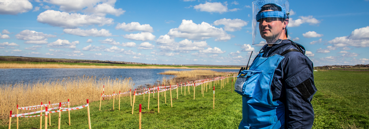 A HALO Trust deminer stands in a rural minefield next to a lake, with contamination clearly marked in front of him