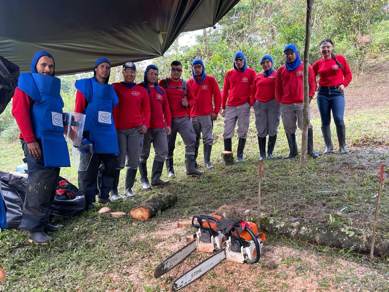 A group of HALO deminers in red shirts and blue PPE stand together under a tarp in the field.