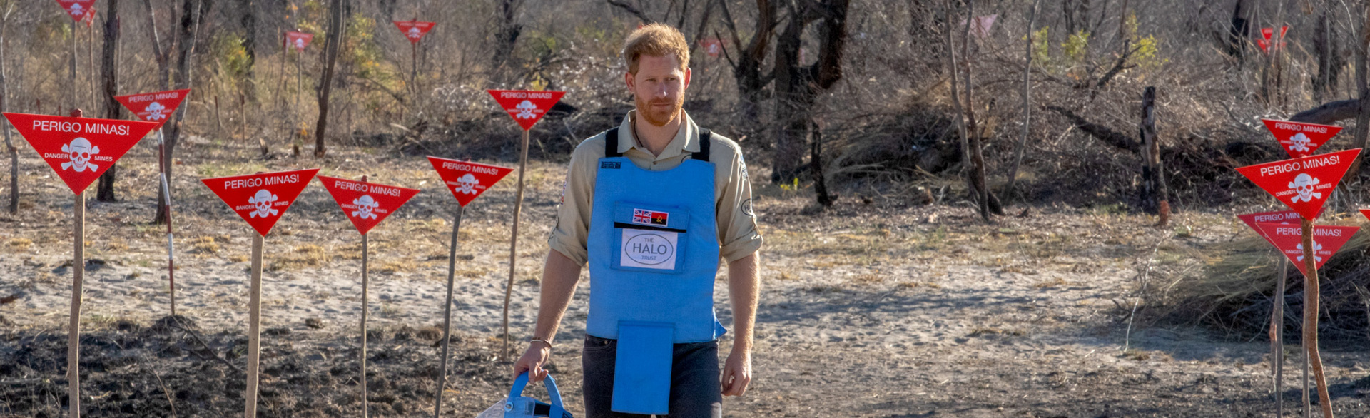 Prince Harry walks through a minefield in Angola, wearing PPE and holding a visor next to red danger signs
