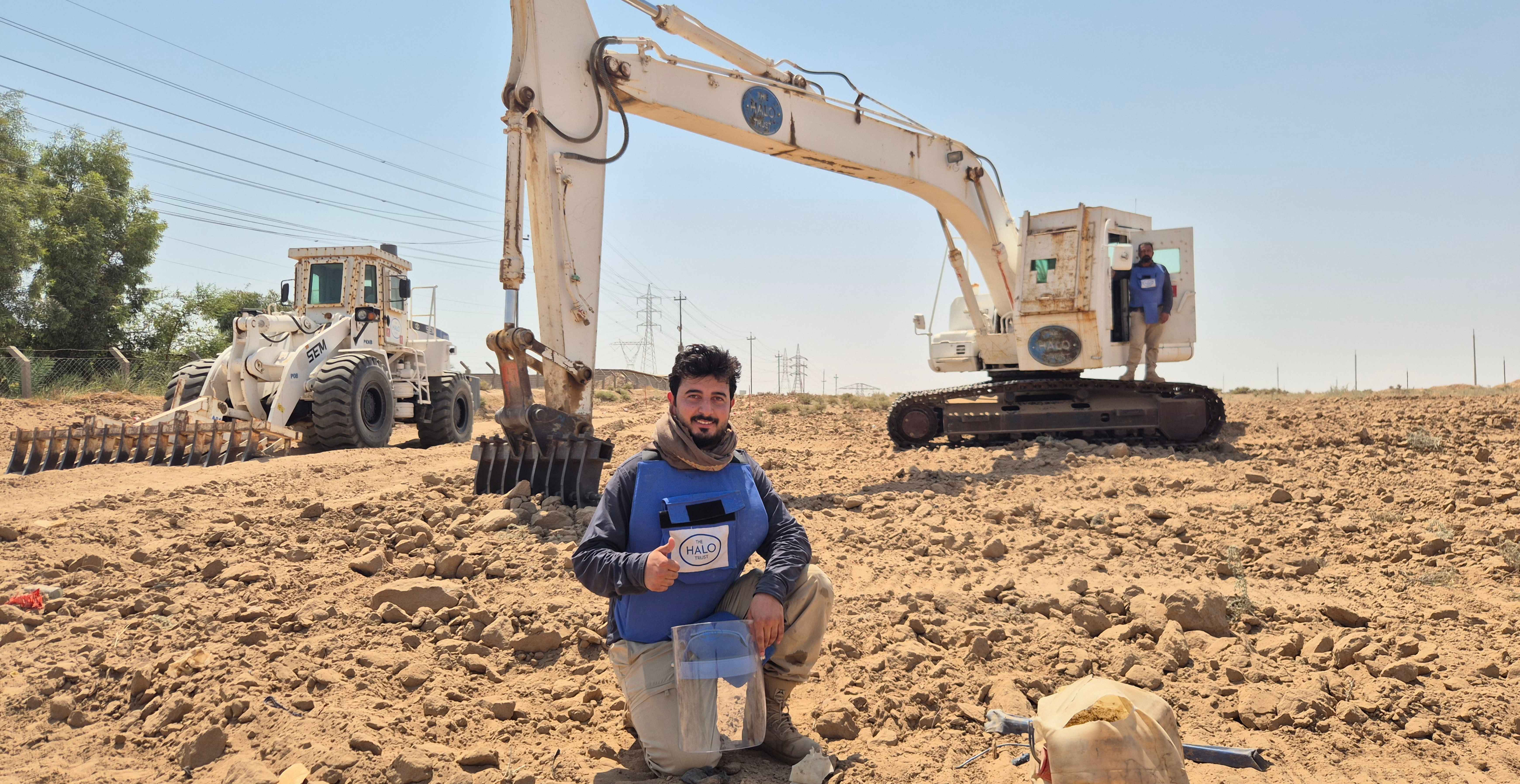 A HALO deminer kneels before digging equipment on a minefield and smiles at the camera