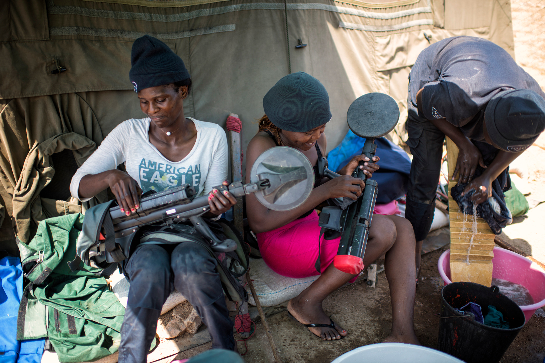 Two women hold demining gear while one washes their clothes outside a camp