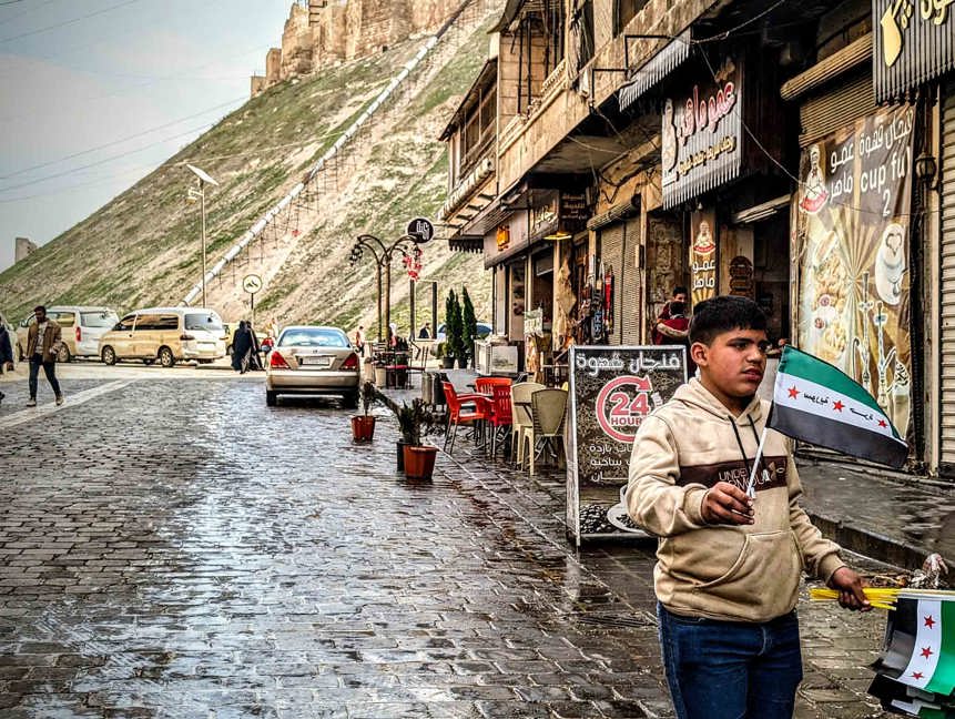 In Aleppo Citadel a boy stands by the side walk with a Syrian flag