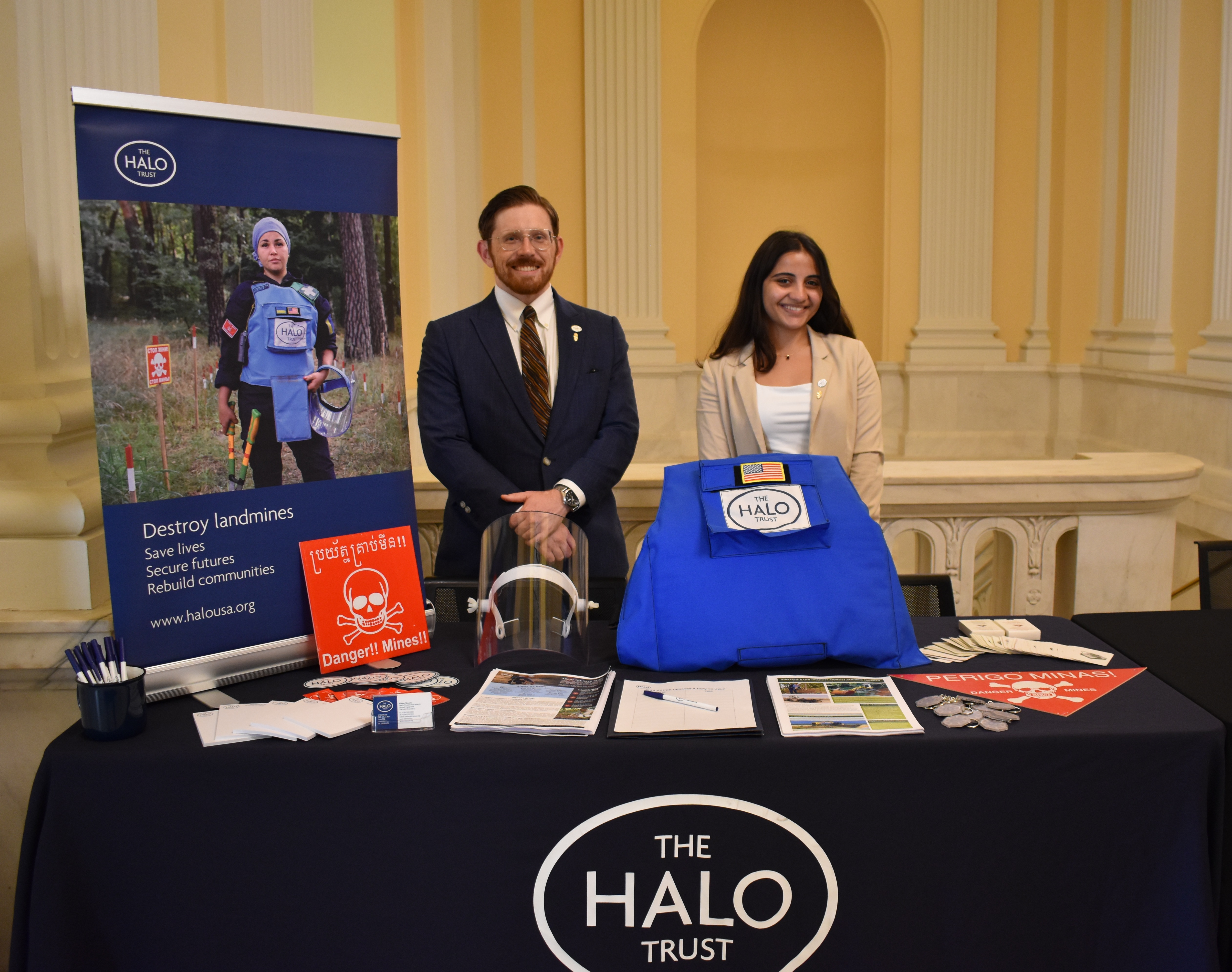 Two HALO staffers smile behind a demining equipment display table 